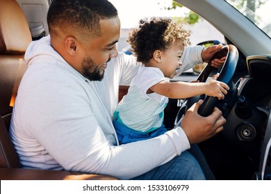 Shot Of A Little Boy Sitting On His Father's Lap In A Car And Learning To Drive. Young Parent Playing With Son In Car.