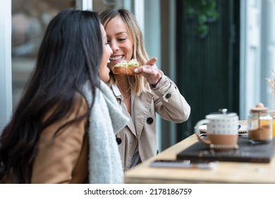 Shot Of Lovely Girls Couple Sharing A Brunch Together While One Of Them Feeds The Other With Avocado Toast On The Healthy Coffee Shop Terrace.