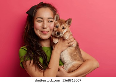 Shot Of Lovely Korean Girl Being In Love With Her Shiba Inu Dog, Embraces Pet With Smile, Has Dark Hair, Wears Green T Shirt, Poses With Animal Against Pink Background. Display Of Affection.