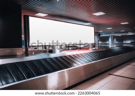 Shot in low-key of a very large rectangular blank billboard mockup facing a baggage claim in an airport terminal; a big white blank advertisement template indoors in front of a baggage handling system