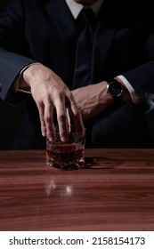 Shot of male hands holding a glass in the form of a spherical ice. There is a designer transparent asymmetrical glass with a thick bottom on a brown striped table. The cup is filled with whiskey.