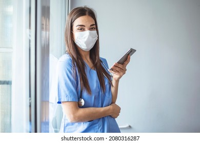 Shot Of A Masked Young Doctor Using A Smartphone. Sad Modern Physician Woman Using Phone App. Female Nurse Using Smartphone On Her Way To Work. Taking Break, Nurse Talks On Smart Phone