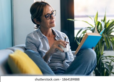 Shot of mature beautiful woman reading a book while drinking a cup of coffee sitting on couch at home. - Powered by Shutterstock