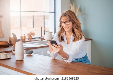 Shot of a mature woman using a mobile phone and drinki tea while standing in the kitchen at home. - Powered by Shutterstock