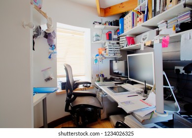 A shot of a messy desk in a home office, the room is small and cluttered, on the desk is three computer monitors and office supplies.