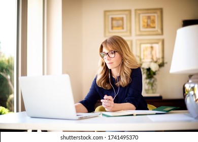 Shot of middle aged businesswoman using earphone while sitting behind her laptop and having discussion and online meeting in video call. Businesswoman working from home. Home office.