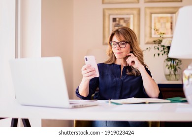 Shot of middle aged woman sitting at desk while working and text messaging. Home office. 