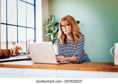 Shot of middle aged woman using her laptop while working in the kitchen from home. Home office.