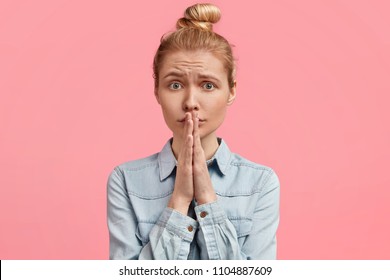 Shot of miserable young female keeps palms together near mouth, has pleading expression, light hair combed in bun, wears denim fashionable jacket, poses against pink background. Please, help me!