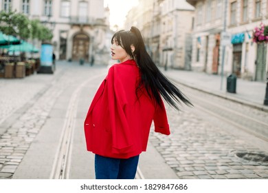 A Shot In The Motion Of Charming Attractive Young Asian Woman, Wearing Red Blazer And Jeans, Walking In The Old European City. Outdoor City Portrait Of Fashion Woman Model