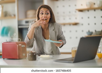 Shot Of A Multi-tasking Young Business Woman Having A Breakfast And Using Laptop In Her Kitchen While Getting Ready To Go To Work.