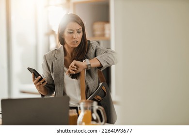Shot of a multi-tasking young business woman looking on watch and using smartphone in her kitchen while getting ready to go to work. - Shutterstock ID 2264093677