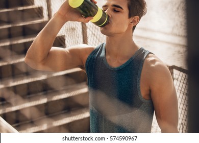 Shot of muscular young man drinking water by the beach after workout. Runner drinking water after exercising and taking break.