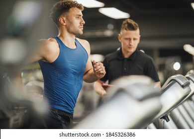 Shot of an muscular young man jogging on the treadmill at the gym. He is working out with personal trainer. - Powered by Shutterstock