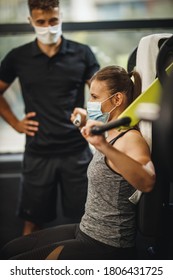 Shot Of A Muscular Young Woman With Protective Mask Working Out With Personal Trainer At The Gym Machine During Covid-19 Pandemic. She Is Pumping Up Her Shoulder Muscule With Heavy Weight.