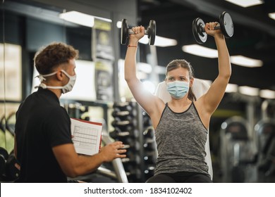 Shot Of A Muscular Young Woman With Protective Mask Working Out With Personal Trainer At The Gym During Covid-19 Pandemic. She Is Pumping Up Her Muscule With Dumbbell.