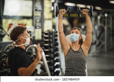 Shot of a muscular young woman with protective mask working out with personal trainer at the gym during Covid-19 pandemic. She is pumping up her muscule with dumbbell.
