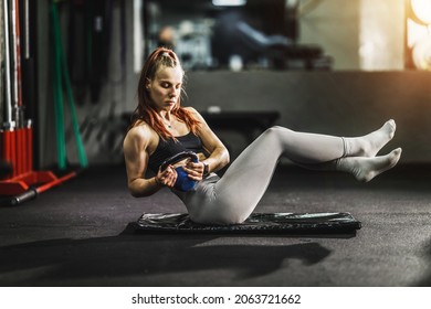 Shot of a muscular young woman in sportswear doing sit ups with kettlebell while exercising in the gym.