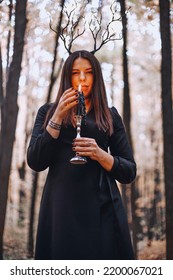 Shot Of Mysterious Woman In Black Dress Holding Candle On Candlestick In Her Hands Performing Magic Ritual In Dark Autumn Forest. Witchcraft Concept