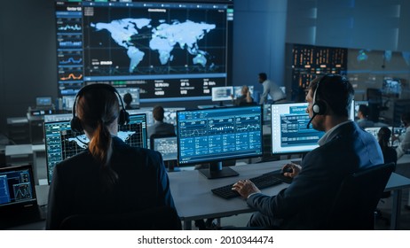 Shot Of Officers In A Surveillance Control Center With Police Global Map Tracking On A Big Digital Screen. Monitoring Room Employees Sit In Front Of Computer Displays And Analyze Big Data.