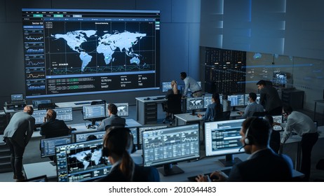 Shot Of Officers In A Surveillance Control Center With Police Global Map Tracking On A Big Digital Screen. Monitoring Room Employees Sit In Front Of Computer Displays And Analyze Big Data.