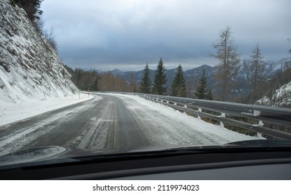 A Shot On The Move From Behind The Windshield Of An Electric Car With Snow-covered Mountains. Cold Cloudy Winter Day. POV First Person View Shot On A Snowed Mountain Road. Selective Focus.