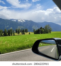 A shot on the move from the driver window at green fields and snowed mountains. Sunny spring day. POV first person view shot on a plain road in the morning. Selective focus.
