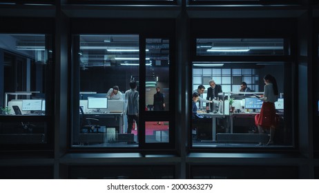 Shot From Outside The Window: Businessmen And Businesswomen Working In The Office. Managers And Specialists Doing Financial Business In The Evening. Employees Work On Computers And Delegate Tasks.