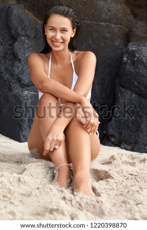 Shot of pleased tanned young female model has slender legs, slim figure, poses on stony shore beside dark crag, recreats at remote sandy beach, has toothy smile, being wet after swimming in sea.