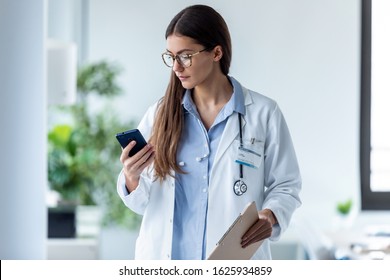 Shot Of Pretty Female Doctor Using Her Mobile Phone While Standing In Medical Consultation.