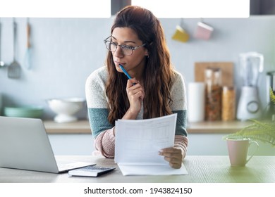 Shot of pretty young business woman working with computer while consulting some invoices and documents in the kitchen at home. - Shutterstock ID 1943824150