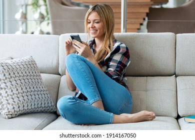 Shot Of Pretty Young Smiling Woman Using Mobile Phone Sitting On A Couch At Home.