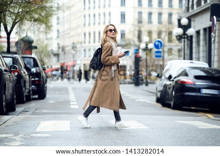 Shot of pretty young woman crossing the street while holding the smartphone and looking sideways.