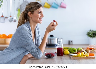 Shot of pretty young woman eating red berries while sitting in the kitchen at home.