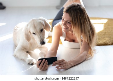 Shot Of Pretty Young Woman With Her Dog Using Mobile Phone While Lying On The Floor At Home.