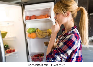 Shot of pretty young woman hesitant to eat in front of the fridge in the kitchen.