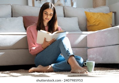 Shot of pretty young woman reading a book and drinking a cup of coffee while sitting on the floor at home. - Powered by Shutterstock