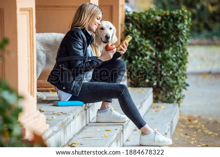 Shot of pretty young woman sending messages with her smart phone while sitting with her lovely golden retriever dog in the park in autumn.