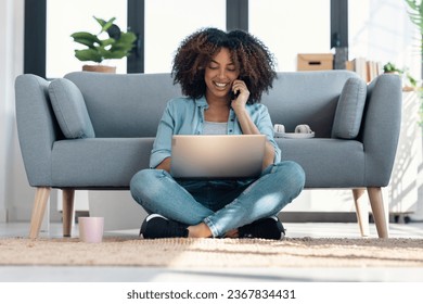 Shot of pretty young woman talking with smartphone while working with laptop sitting on the floor at home. - Powered by Shutterstock