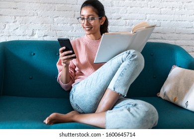 Shot Of Pretty Young Woman Using Her Mobile Phone While Reading A Book On Sofa At Home.