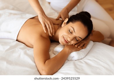 Shot of relaxed brunette lady with eyes closed in contentment lying during relaxing therapeutic back massage in modern spa setting. Professional masseur massaging client's neck and shoulders - Powered by Shutterstock