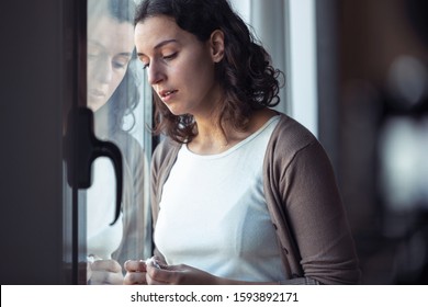 Shot Of Sad Young Woman Crying While Looking Through The Window At Home.