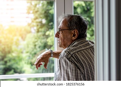 Shot of a senior man looking through window of his apartment. Portrait of a elder man in contemplation near window looking away and thinking. Pensive grey hair senior standing and relaxing, copy space