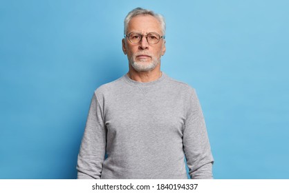 Shot of serious bearded mature European man dressed in casual grey jumper looks directly at camera being confident in himself poses against blue studio background. Handsome grandfather on pension