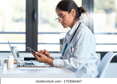 Shot of serious female doctor using her smartphone while working with laptop in the consultation.