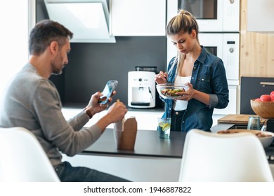 Shot Of Serious And Unhappy Young Couple Eating While Using Smartphone In The Kitchen At Home.