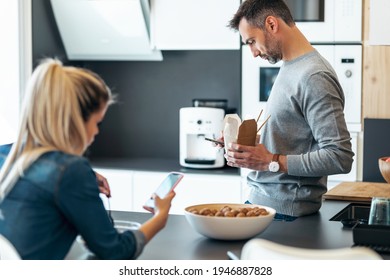 Shot Of Serious And Unhappy Young Couple Eating While Using Smartphone In The Kitchen At Home.