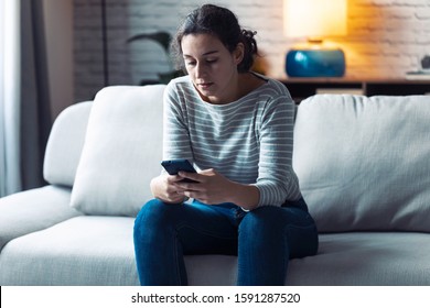 Shot Of Serious Young Woman Using Her Mobile Phone While Sitting On Sofa In The Living Room At Home.