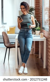 Shot Of Smart Business Woman Using Mobile Phone While Drinking A Cup Of Coffee Leaning On The Table In Living Room At Home.