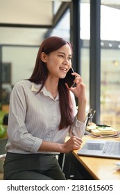 Shot Of Smiling Asian Woman Office Worker Talking On Mobile Phone While Sitting At Wooden Counter At Office